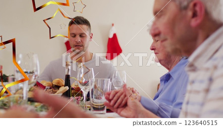 Image of stars over caucasian family with santa hats praying before christmas dinner 123640515