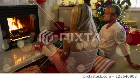 Image of dots moving over caucasian couple sitting by fireplace under christmas tree 123640908