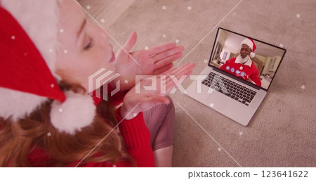 Image of snow falling over happy caucasian woman in santa hat on laptop image call with family 123641622