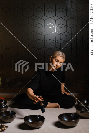 Woman practicing sound healing with singing bowls in a tranquil indoor space decorated with plants and soft lighting during a peaceful evening session 123642330