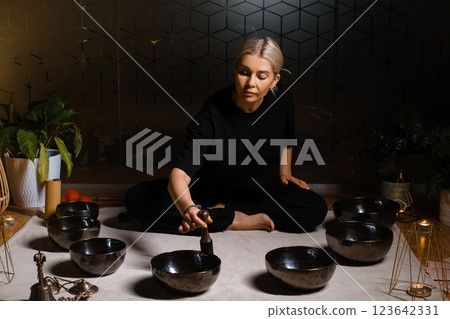 Woman practicing sound healing with singing bowls in a tranquil indoor space decorated with plants and soft lighting during a peaceful evening session 123642331