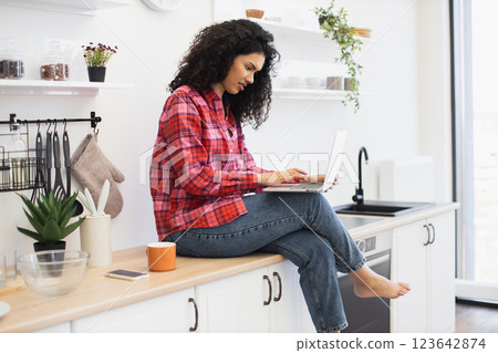 Young African woman wearing casual outfit sitting on kitchen counter working on laptop. Relaxing morning atmosphere featuring coffee, kitchen decor, and natural indoor light for productivity. Young African woman wearing casual outfit sitting on kitchen counter working on laptop. Relaxing morning atmosphere featuring coffee, kitchen decor, and natural indoor light for productivity. 123642874