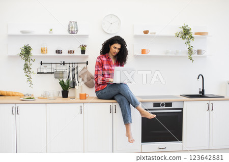 Young African adult woman wearing casual clothes using laptop in modern white kitchen interior 123642881
