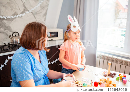 Wide shot of Caucasian mother and her little daughter are drinking tea together in the kitchen. Little girl put on a homemade paper rabbit mask. Concept of the Easter Spring festival 123648036