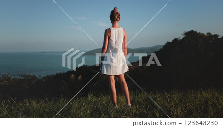 Woman in a white dress stands on a grassy hill, enjoying a tropical island view at sunset. The scene is serene and beautiful, evoking summer vacation vibes Woman in a white dress stands on a grassy hill, enjoying a tropical island view at sunset. The scene is serene and beautiful, evoking summer vacation vibes 123648230