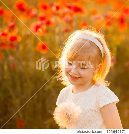 A smiling blonde girl blows a large white dandelion with a poppy field and sunset in the background A smiling blonde girl blows a large white dandelion with a poppy field and sunset in the background 123649171