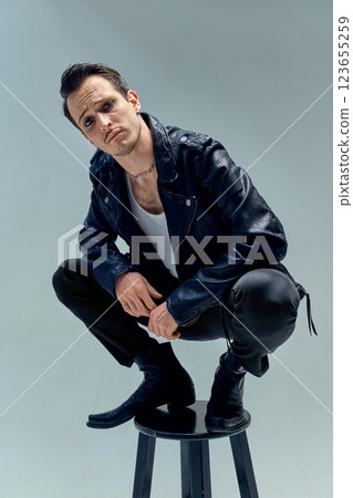 Edgy young man, performer crouching on stool, piercing gaze locked in intensity against soft-toned background. Concept of music Edgy young man, performer crouching on stool, piercing gaze locked in intensity against soft-toned background. Concept of music 123655259
