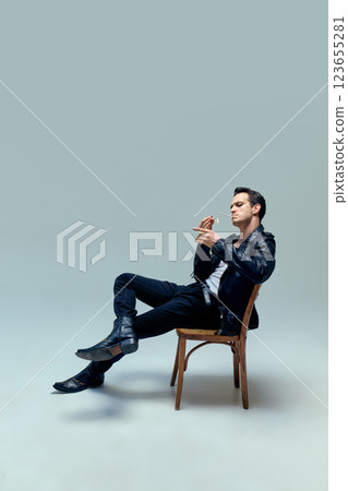 Man dressed vintage attire lounging effortlessly, cigarette lit with timeless confidence, embodying essence of cinematic cool against studio background. Man dressed vintage attire lounging effortlessly, cigarette lit with timeless confidence, embodying essence of cinematic cool against studio background. 123655281