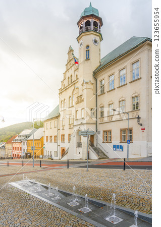 Visitors admire the elegant architecture of Jachymov's town hall. The building features a striking clock tower and is surrounded by charming streets and decorative fountains. Visitors admire the elegant architecture of Jachymov's town hall. The building features a striking clock tower and is surrounded by charming streets and decorative fountains. 123655954