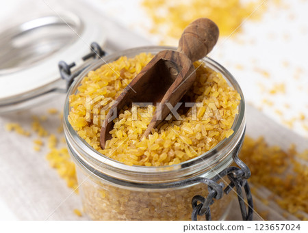 Glass jar of raw dry uncooked yellow bulgur grain with a wooden scoop on napkin closeup 123657024