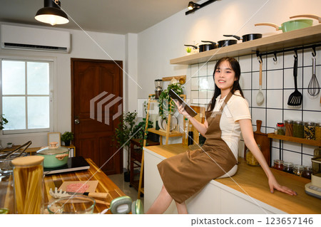 Cheerful young woman wearing an apron sitting on the kitchen counter and using digital tablet 123657146