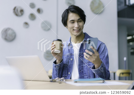 一名年輕男子一邊在電腦前工作,一邊拿著智慧型手機和咖啡。 一名年輕男子一邊在電腦前工作,一邊拿著智慧型手機和咖啡。 123659092