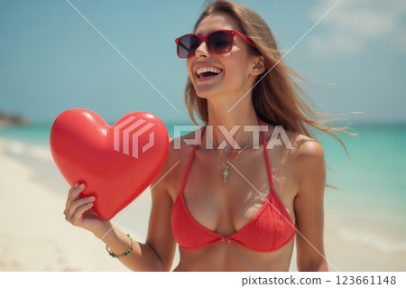 portrait of young beautiful girl wearing bikini swimsuit and posing on resort beach with view of sea and bright sky. She holds a heart shaped valentine in her hands 123661148