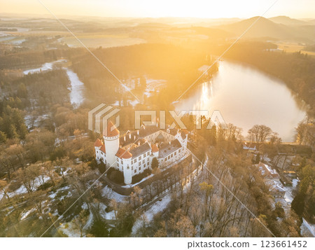 Konopiste Castle stands majestically surrounded by lush nature, with soft sunlight illuminating its structure. The tranquil lake reflects the warm hues of the setting sun, enhancing the scenic beauty. 123661452
