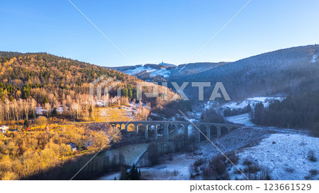 The railway stone bridge in Novina is illuminated by a winter sunset, with the majestic Jested Mountain in the background, surrounded by serene nature and frosty landscapes. 123661529