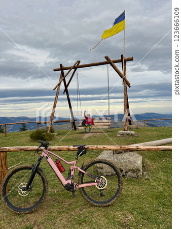 Pink electric mountain bike in the foreground. Person resting from biking on large wooden swing structure in the background. Carpathians, Ukraine. Pink electric mountain bike in the foreground. Person resting from biking on large wooden swing structure in the background. Carpathians, Ukraine. 123666109