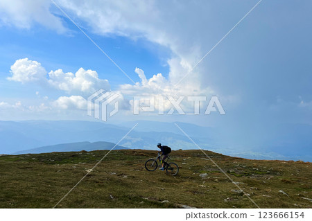 Cyclist man riding electric mountain bike outdoors. Male tourist biking along grassy trail in the mountains, wearing helmet and backpack. Concept of sport, active leisure and nature. 123666154