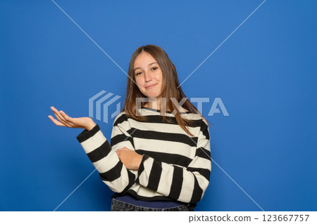 Beautiful Hispanic teenage girl about 15 years old wearing a striped sweater holds her hand up with blank copy space to the side. Isolated on blue studio background. Beautiful Hispanic teenage girl about 15 years old wearing a striped sweater holds her hand up with blank copy space to the side. Isolated on blue studio background. 123667757