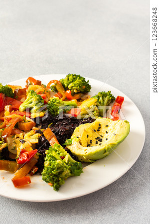 A hearty and nutritious breakfast of black rice with stewed vegetables like tomatoes, zucchini, peppers, and onions, fresh salad with tomatoes, broccoli, and radish, avocado 123668248
