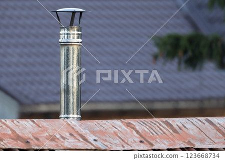 A metallic chimney pipe with a cap stands on a red roof 123668734