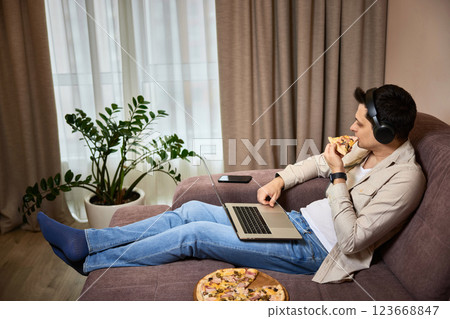 Young man sitting on sofa and eating pizza while watching movie on laptop Young man sitting on sofa and eating pizza while watching movie on laptop 123668847