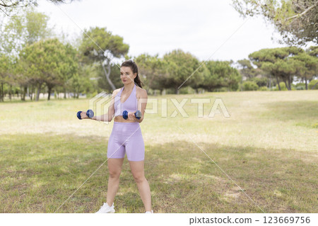 A Young Woman, in the Midst of an Outdoor Workout in the Park, Lifting Weights and Enhancing Her Physical Strength 123669756
