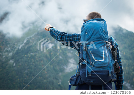 A hiker with a blue expedition backpack and plaid sweatshirt points to misty mountain peaks. Back view. Cloud-shrouded alpine landscape and valleys in front of him Mountain hiking A hiker with a blue expedition backpack and plaid sweatshirt points to misty mountain peaks. Back view. Cloud-shrouded alpine landscape and valleys in front of him Mountain hiking 123670106