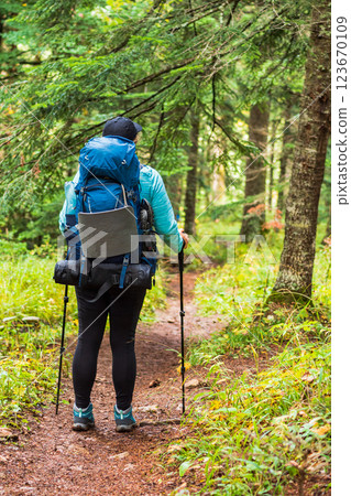 A girl hiker with a large blue backpack is walking along a winding forest path. In her hands are trekking poles. The scene evokes a sense of adventure and oneness with nature 123670109