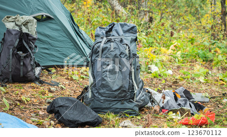 A large gray hiking backpack stands in the foreground, next to a partially visible green tent. Other hiking gear is scattered around, including the backpack and rain cover. Mountain Hiking A large gray hiking backpack stands in the foreground, next to a partially visible green tent. Other hiking gear is scattered around, including the backpack and rain cover. Mountain Hiking 123670112