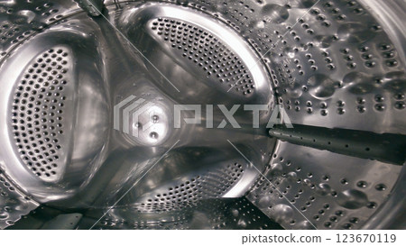 A close-up of the inside of a washing machine drum, showing the perforated metal surface and the central agitator with three arms. The drum is made of stainless steel and has a reflective surface A close-up of the inside of a washing machine drum, showing the perforated metal surface and the central agitator with three arms. The drum is made of stainless steel and has a reflective surface 123670119
