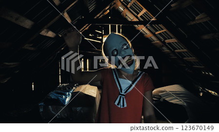 A young girl wearing a creepy mask with big eyes and a wide smile stands in a dark, shadow-covered attic. She waves her right hand. Close-up of a child monster 123670149
