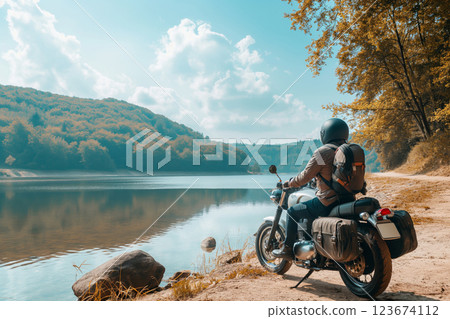 Man in full gear pauses on his motorcycle by a tranquil lake, reflecting on the journey amidst autumn's golden hues 123674112