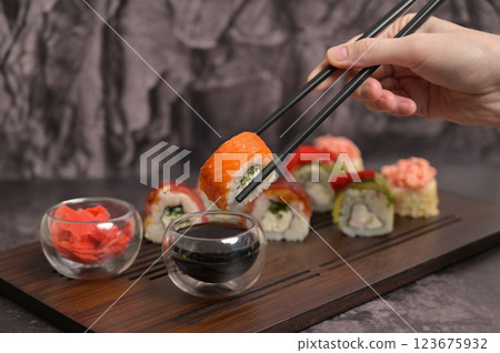Woman's hand holding sushi roll used chopsticks and dipping in soy sauce. Set of sushi roll with rice and fish on wooden serving board. Dark black texture background. Close up. Japan menu. Asian food 123675932