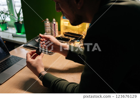 Focused technician repairing smartwatch battery using tweezers for precise component extraction and replacement in dark workshop sitting at desk with laptop computer. 123676129