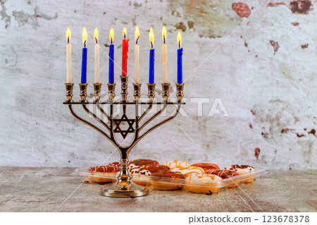 Beautifully lit menorah featuring blue red candles stands next to an assortment of traditional sweets, capturing festive spirit of Hanukkah on rustic table. 123678378