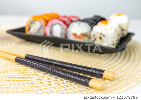 Variety of sushi rolls on black plate with chopsticks on woven mat Variety of sushi rolls on black plate with chopsticks on woven mat 123679789