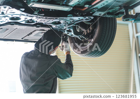 一名男性機械師/維修技師正在對一輛被抬起的汽車底部進行工作、檢查和維修 一名男性機械師/維修技師正在對一輛被抬起的汽車底部進行工作、檢查和維修 123681955