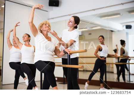 European woman performs an exercise near a ballet barre, where the choreographer helps to do it correctly European woman performs an exercise near a ballet barre, where the choreographer helps to do it correctly 123683754