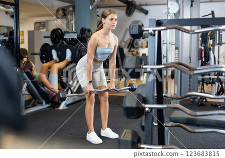 Slender girl trains muscles of arms and back using barbell and differentiated weighting in gym 123683815