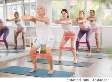 Group of women standing on mats and doing stretch exercises with resistance bands in gym 123683868