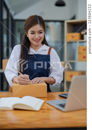 Young woman engaged in online business working with using laptop computer, preparing package for shipping and taking notes customer address at box 123684153