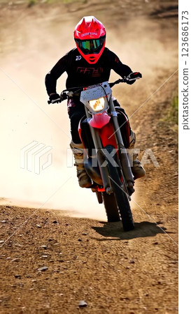 Motocross racer wearing protective equipment balancing on rear wheel, launching over dusty terrain with high-energy dynamic motion, demonstrating professional riding technique 123686173