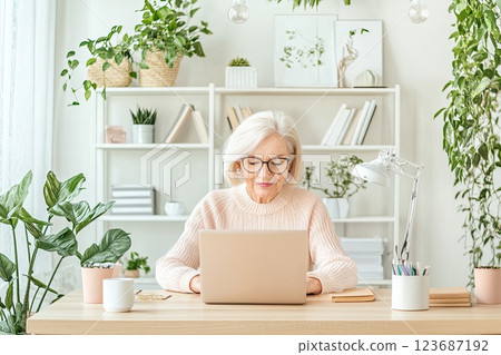 Senior woman working on laptop at home office surrounded by indoor plants during daylight hours Senior woman working on laptop at home office surrounded by indoor plants during daylight hours 123687192