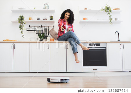 African woman sitting on kitchen counter in modern kitchen controlling robotic vacuum cleaner. Spacious white kitchen environment with shelves, plants, and household appliances. African woman sitting on kitchen counter in modern kitchen controlling robotic vacuum cleaner. Spacious white kitchen environment with shelves, plants, and household appliances. 123687414