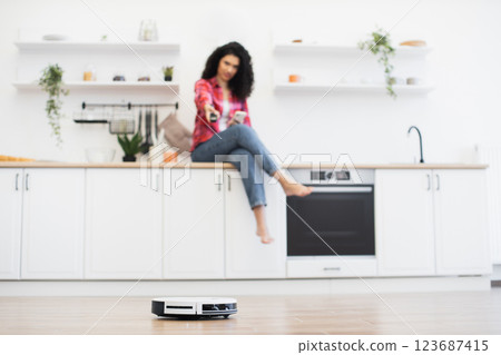 African woman sitting on kitchen counter in modern kitchen controlling robotic vacuum cleaner. Spacious white kitchen environment with shelves, plants, and household appliances. 123687415