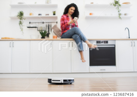 Positive atmosphere modern kitchen African woman sitting counter using smartphone autonomous robot vacuum cleaner operating visible interaction technology convenience lifestyle comfort clean space 123687416