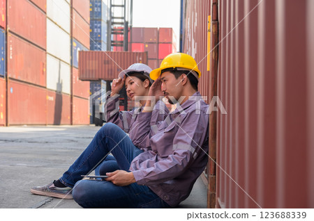 Two Asian man and woman workers in protective helmets take a break while sitting on floor at cargo shipping container yard 123688339
