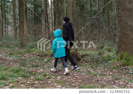 Enjoying a calming walk in the tranquil forest, a child wearing a bright blue jacket strolls alongside an adult in a black hoodie during a cool afternoon 123692552
