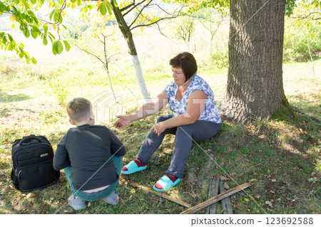 Grandmother and grandson share a moment under a shady tree, enjoying the tranquility of the countryside on a sunny day. Outdoors nature. Grandmother and grandson share a moment under a shady tree, enjoying the tranquility of the countryside on a sunny day. Outdoors nature. 123692588