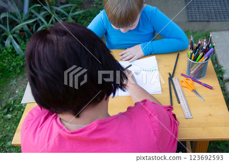 Outdoor art session: A woman guides a child in drawing on a wooden table, surrounded by art supplies, fostering creativity and bonding. 123692593